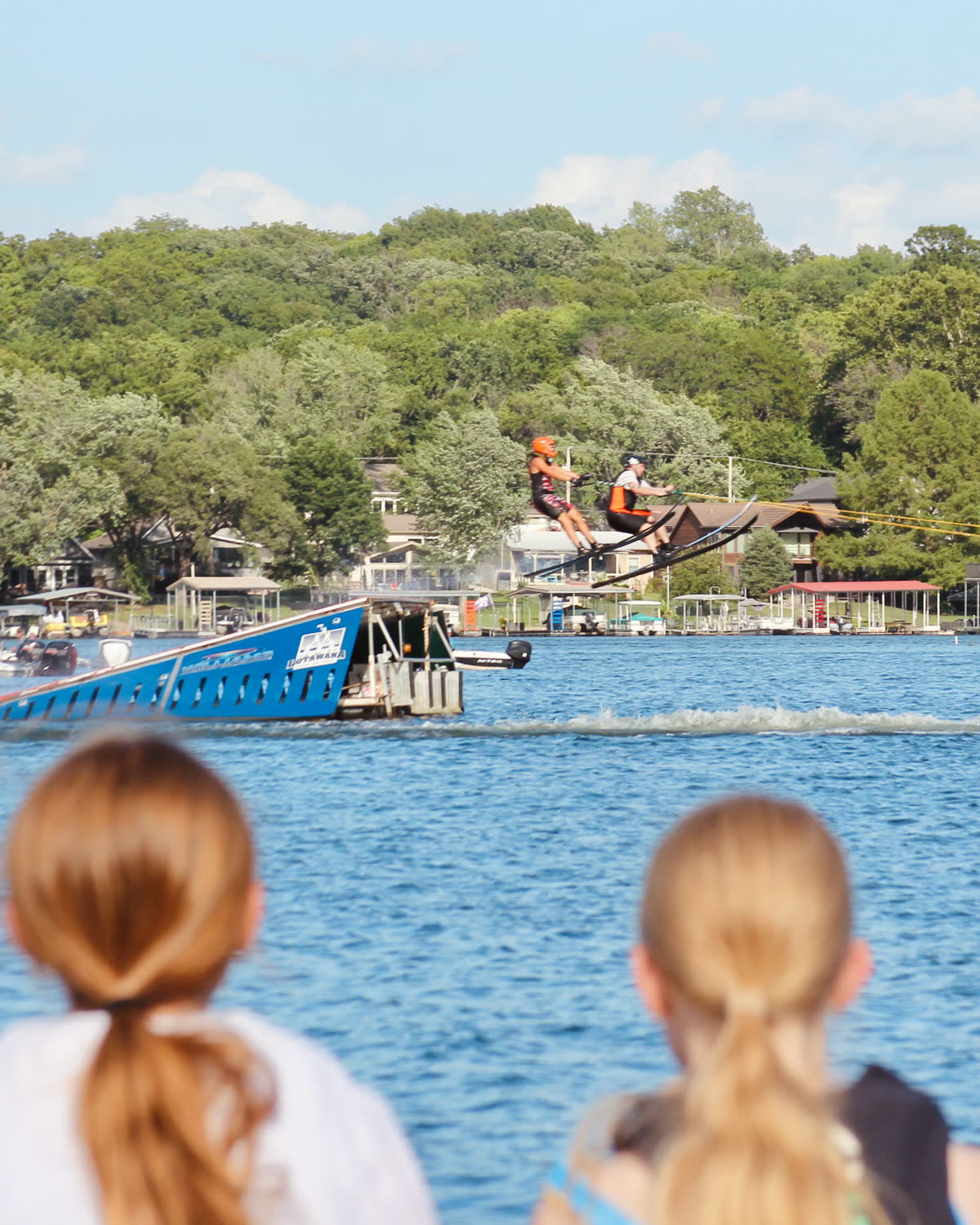 Two kids watching from a boat as water skiers launch off a jump ramp at a Lake Lotawana ski show