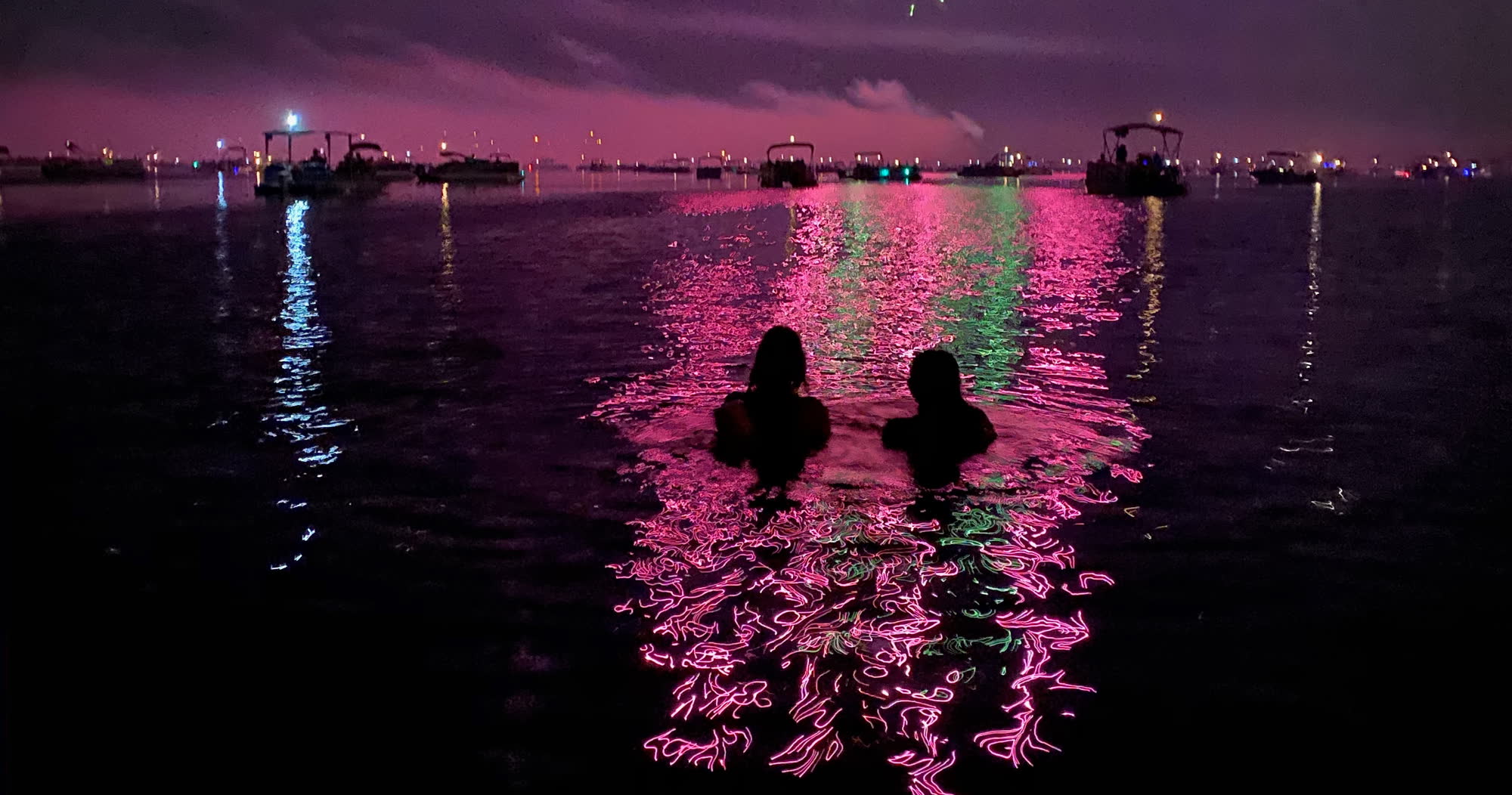 Two swimmers in the lake at night with fireworks reflections streaking pink and green across the water